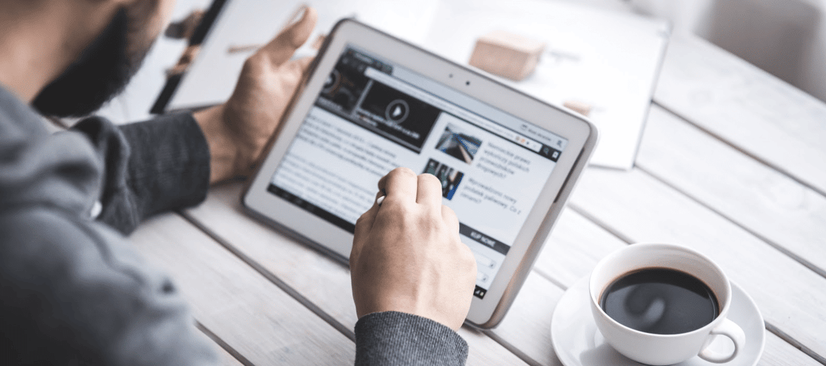 Person tapping a news article on a tablet at a wooden table next to a cup of coffee