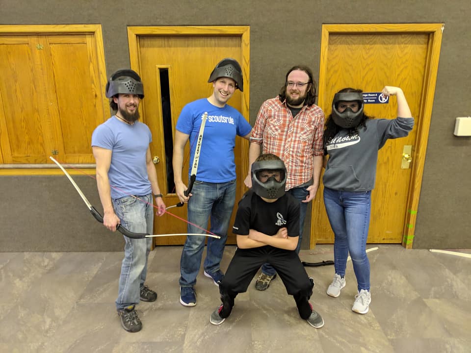 Five Q team members in archery tag masks posing with bows at a team meeting